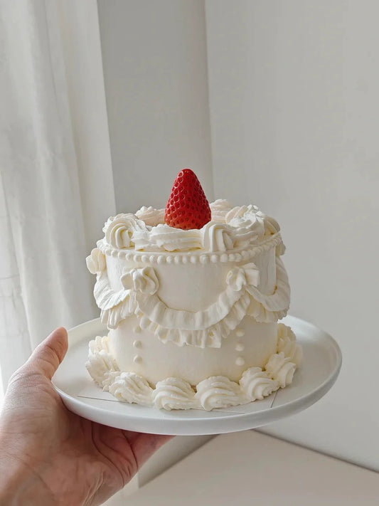 A hand holding a small round white cake decorated with piped frosting borders and topped with a single strawberry, set against a soft neutral indoor background.