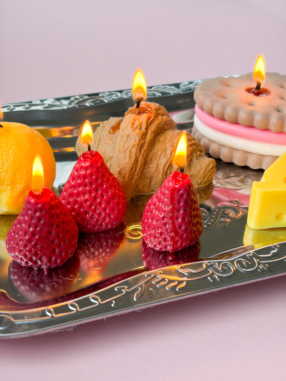 A decorative silver tray holding strawberry-shaped candles, a croissant-shaped candle, a biscuit-shaped candle, a lemon slice, and a wedge of cheese on a soft pink background.