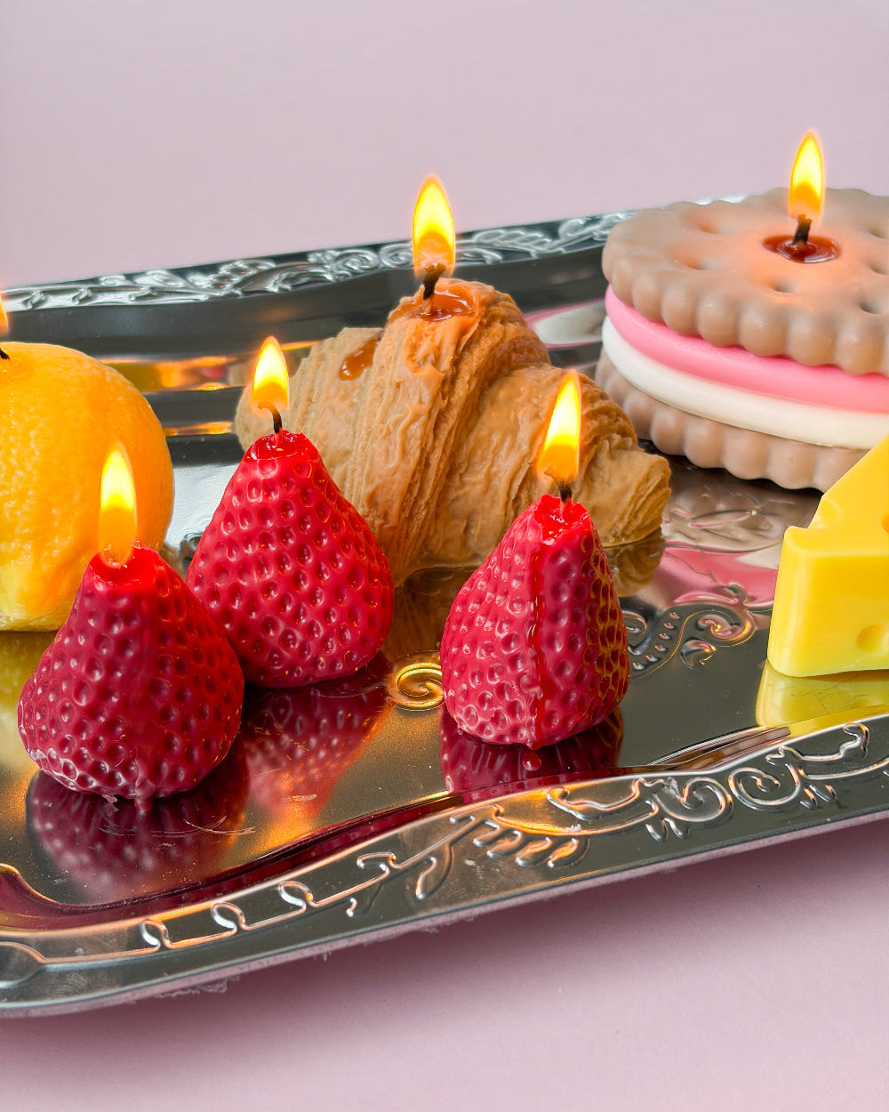 A decorative silver tray holding strawberry-shaped candles, a croissant-shaped candle, a biscuit-shaped candle, a lemon slice, and a wedge of cheese on a soft pink background.