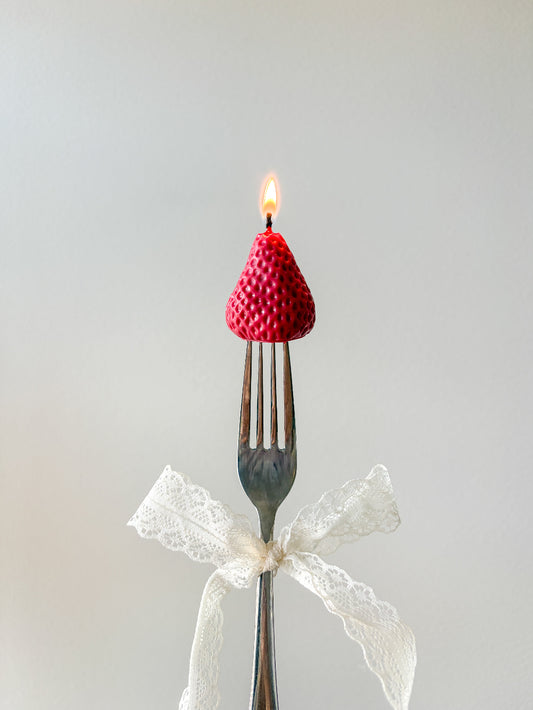 A novelty candle shaped like a red raspberry sits on the tines of a metal fork, with a small flame burning from the top. A white lace ribbon is tied around the handle, and the background is a soft light gray.