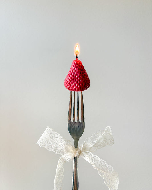 A novelty candle shaped like a red raspberry sits on the tines of a metal fork, with a small flame burning from the top. A white lace ribbon is tied around the handle, and the background is a soft light gray.