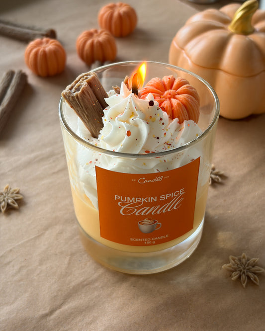 A lit 'Pumpkin Spice Candle' by Candlesticks in a clear glass jar, decorated with whipped cream, a mini pumpkin, cinnamon stick, and orange speckles, set on a brown paper background with autumn pumpkins.