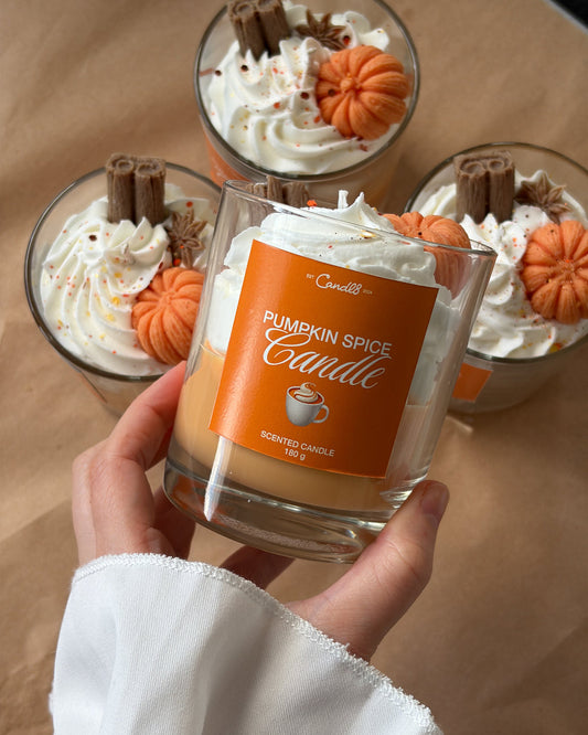 A hand holds a glass jar candle labeled 'Pumpkin Spice Candle' in front of three similar candles topped with whipped cream, orange pumpkin decorations, and cinnamon sticks on a tan surface.