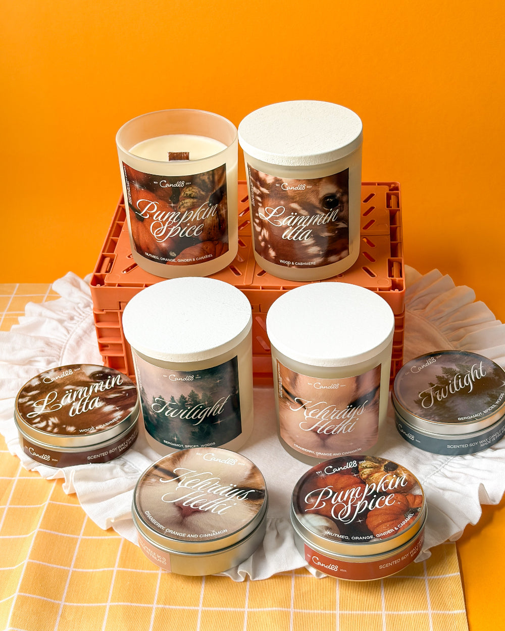 A collection of Candle Couture scented candles in glass jars and tins, featuring labels for Pumpkin Spice, Lammy Ella, Twilight, and Glazed Peach, arranged on a white checkered cloth and orange crate against a solid orange background.