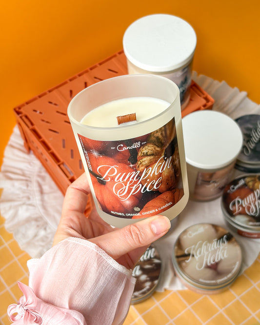 A hand holds a frosted glass candle jar labeled 'Pumpkin Spice' by Candle Co. against a bright orange background, with several other candles and containers arranged around it on a white cloth.