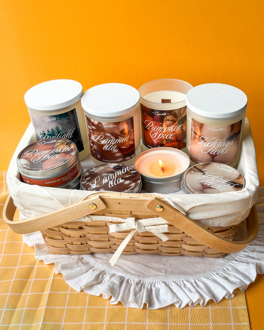 A curated candle gift basket featuring six scented candles in jars and tins, arranged inside a woven wicker basket with a cream fabric liner and wooden handles, set against a solid orange background.