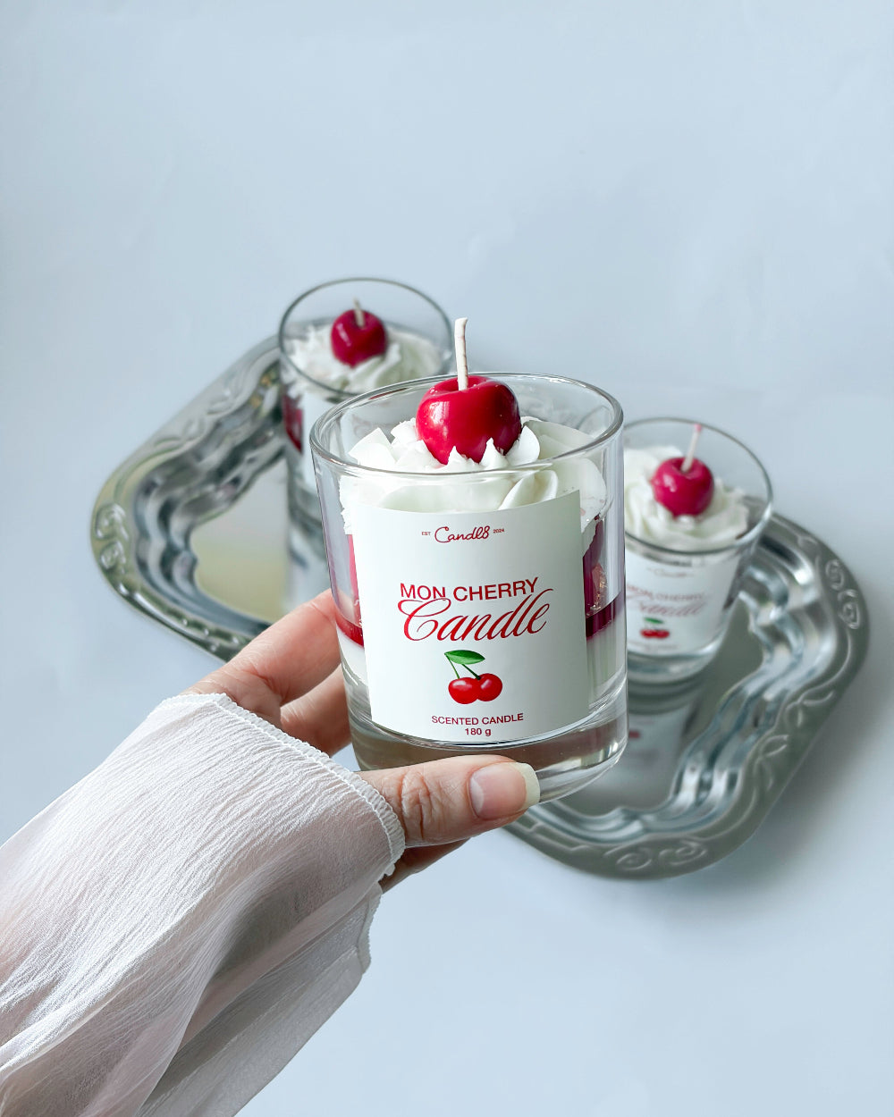 Hand holding a cherry-themed candle with whipped cream and a cherry on top, set against a neutral background.