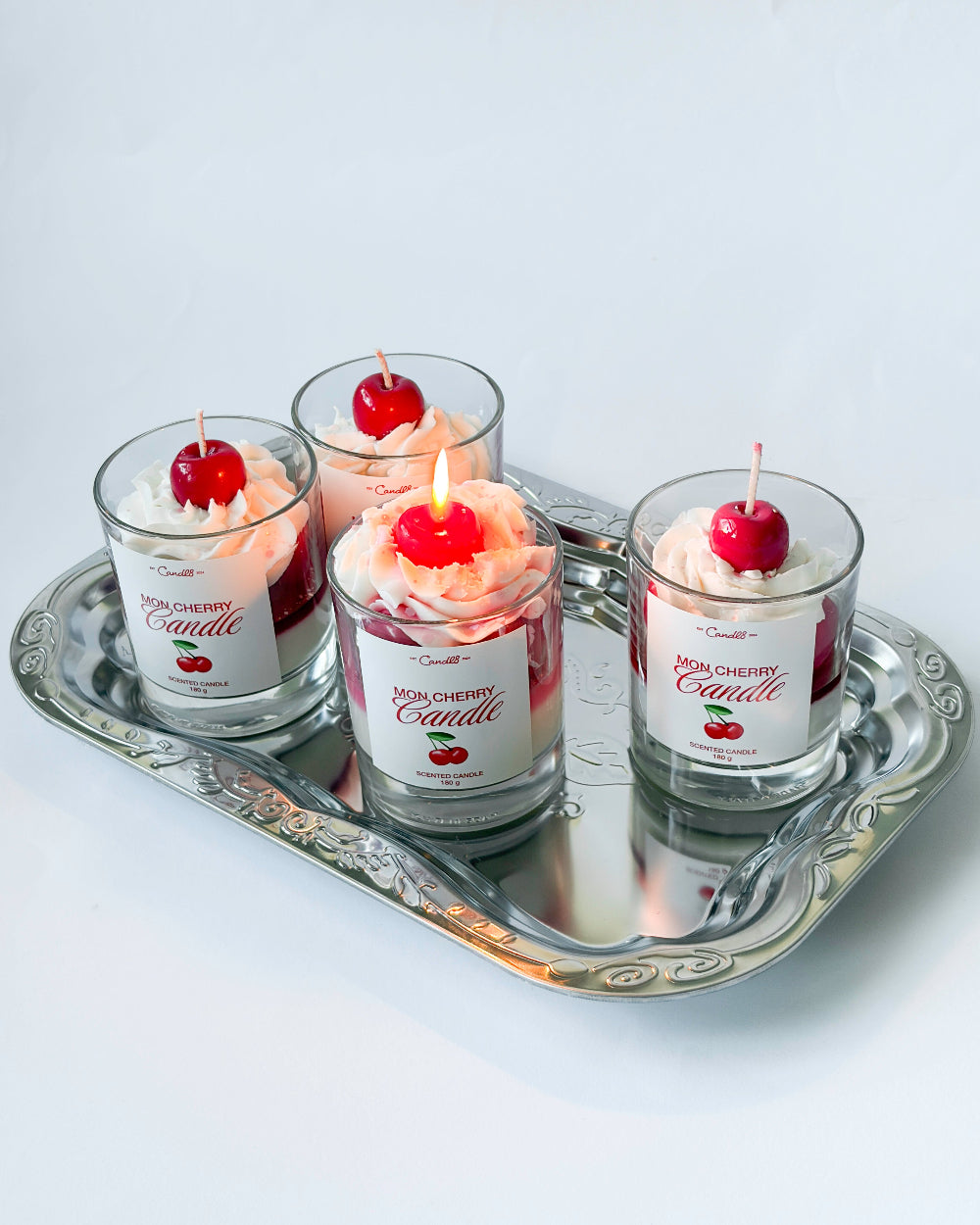 Four small glass jars with cherry on top candles on a silver tray against a white background