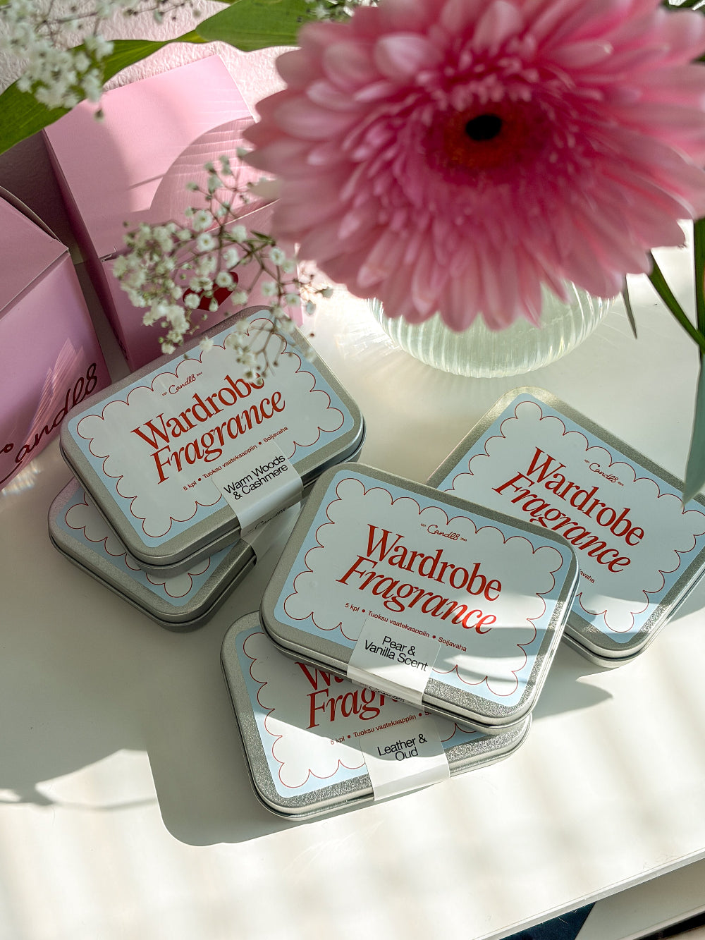 Wardrobe Fragrance tins on a white surface with a pink flower in the background