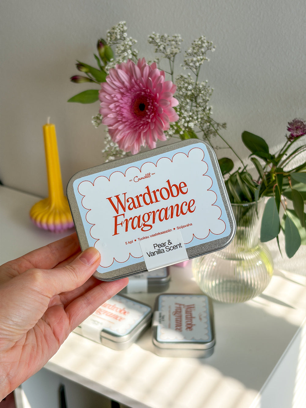 Hand holding a 'Wardrobe Fragrance' tin with flowers and candles in the background