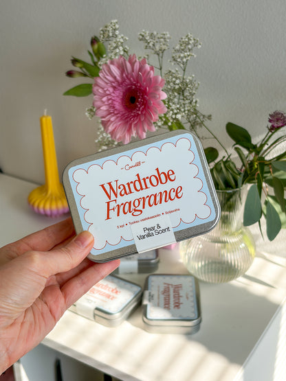 Hand holding a 'Wardrobe Fragrance' tin with flowers and candles in the background