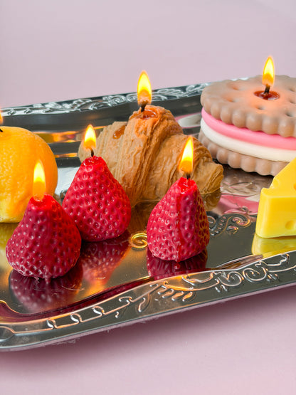 A decorative silver tray holding strawberry-shaped candles, a croissant-shaped candle, a biscuit-shaped candle, a lemon slice, and a wedge of cheese on a soft pink background.