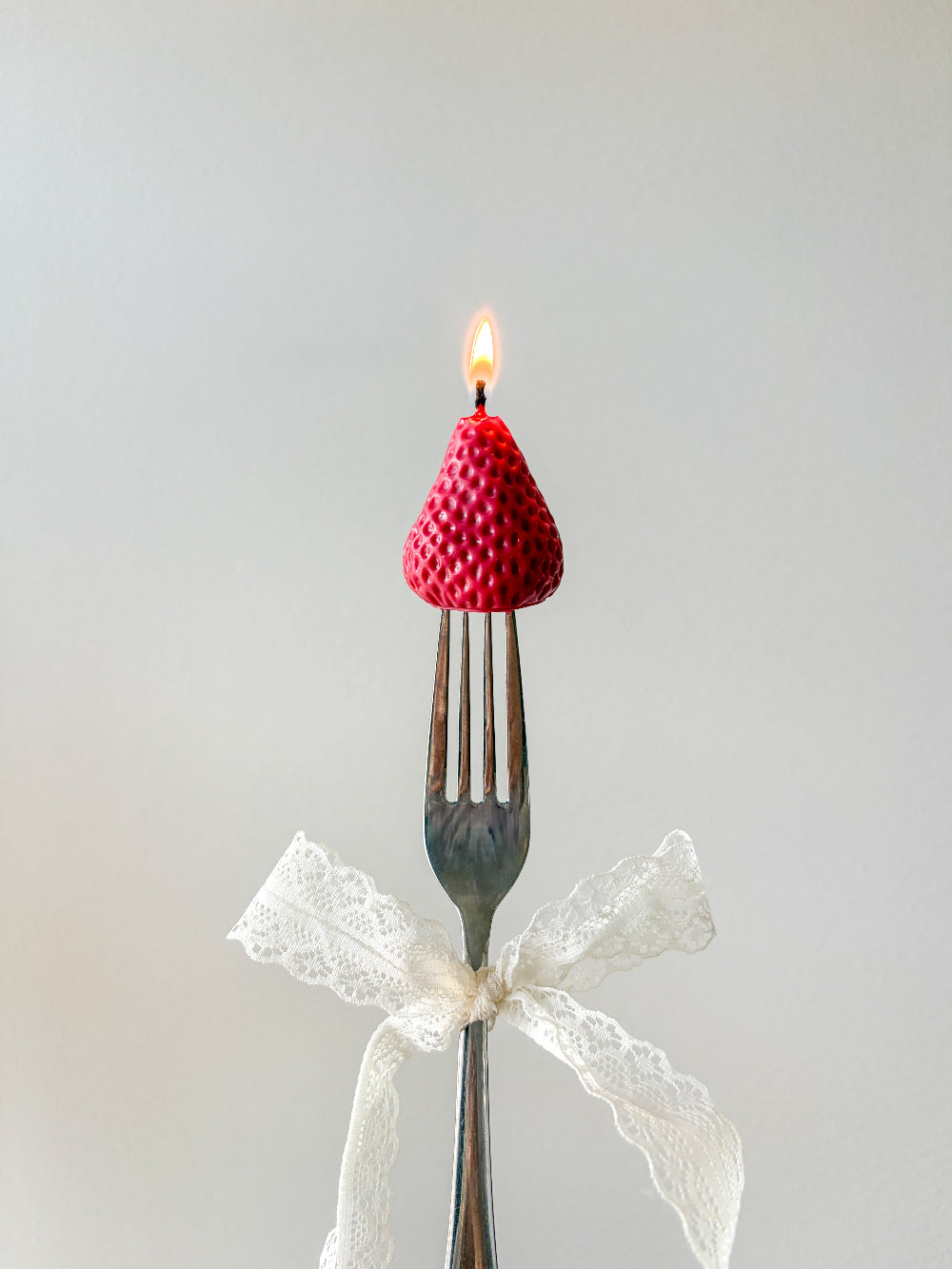 A novelty candle shaped like a red raspberry sits on the tines of a metal fork, with a small flame burning from the top. A white lace ribbon is tied around the handle, and the background is a soft light gray.