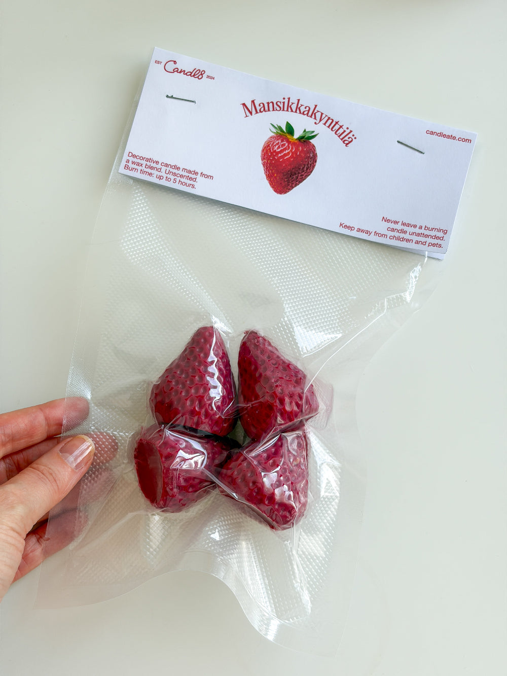 A hand holds a clear plastic vacuum-sealed pouch containing four red strawberry-shaped candies, with a white header card printed with the brand name Candlet and the product name Mansikkakyyntöüli.