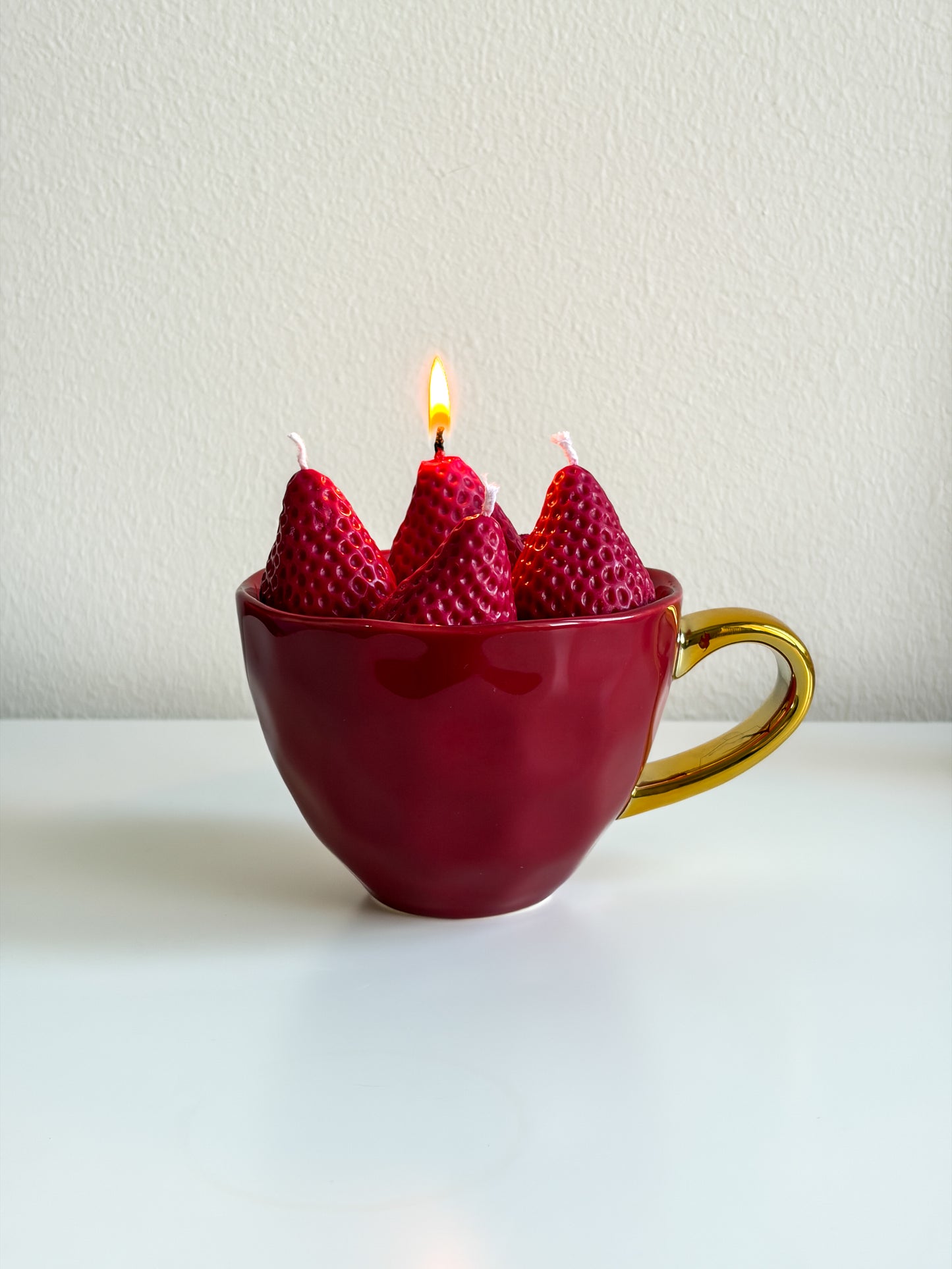 A red ceramic teacup candle holder with a gold handle, containing four strawberry-shaped candles, one of which is lit, set against a plain off-white background.