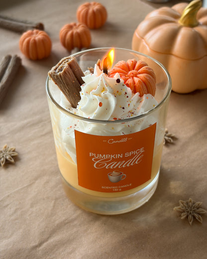 A lit 'Pumpkin Spice Candle' by Candlesticks in a clear glass jar, decorated with whipped cream, a mini pumpkin, cinnamon stick, and orange speckles, set on a brown paper background with autumn pumpkins.