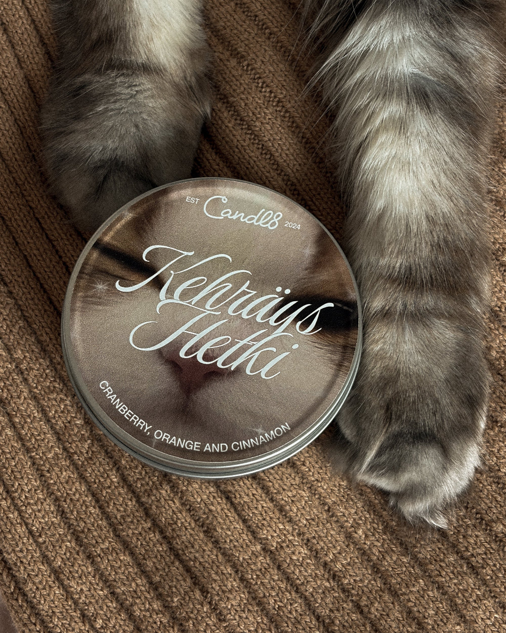A round glass tin of Candell Kebrays Hethki cat perfume, featuring a photo of a cat's face on the label, styled on a brown ribbed knit sweater with a cat's paws resting on top.