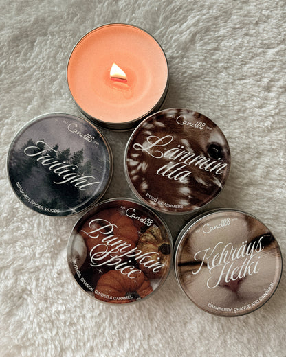 Five round candle tins arranged on a white faux-fur surface, with one tin open and lit in the upper left. The tins feature printed nature-themed labels: Faurial, Lämmintula, Dymman Spice, and Kevlaüs Helki.