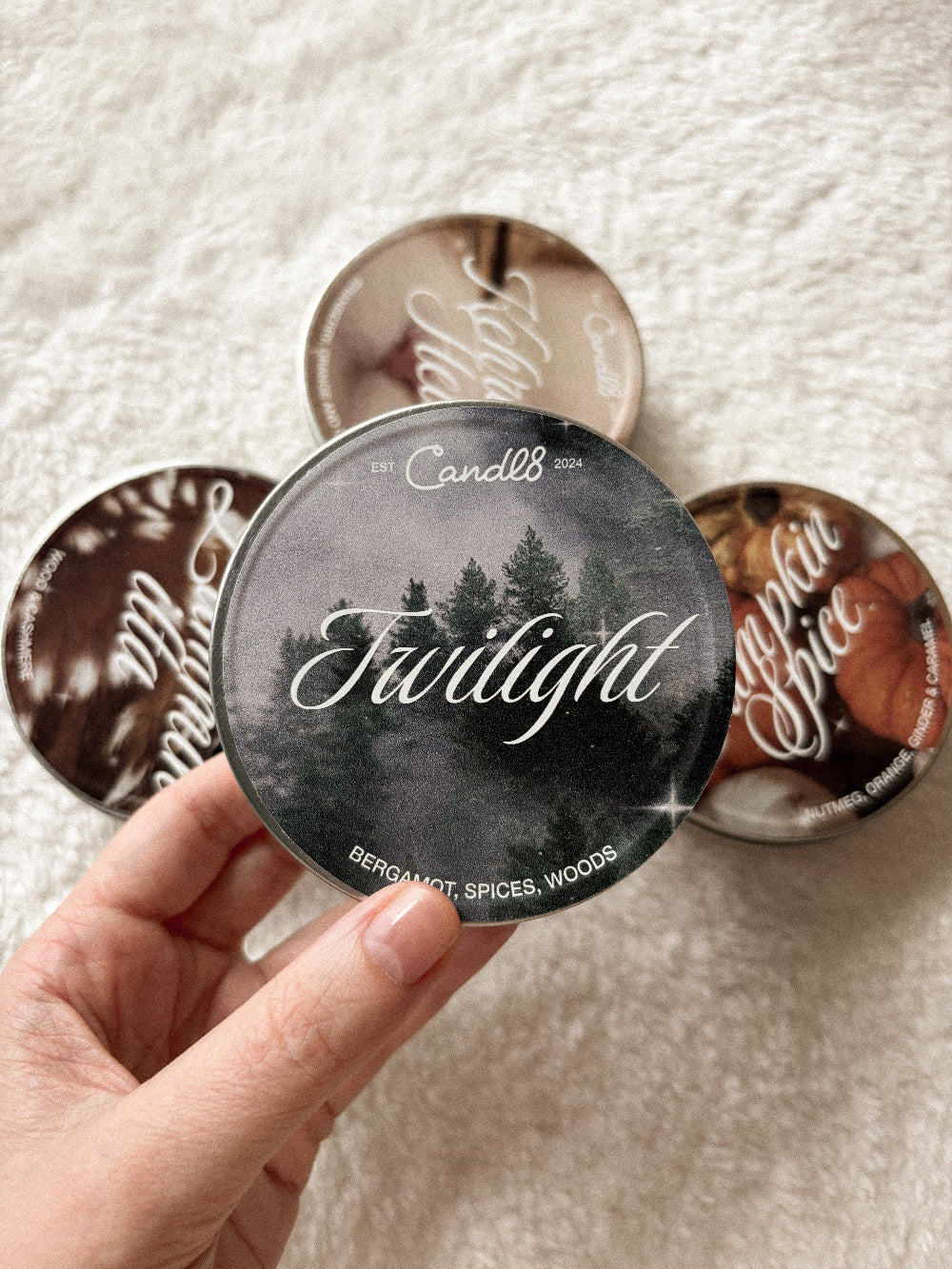 A hand holds a round tin candle labeled 'Twilight' by Candle Co, with three other round candles arranged behind it on a soft white textured fabric background.