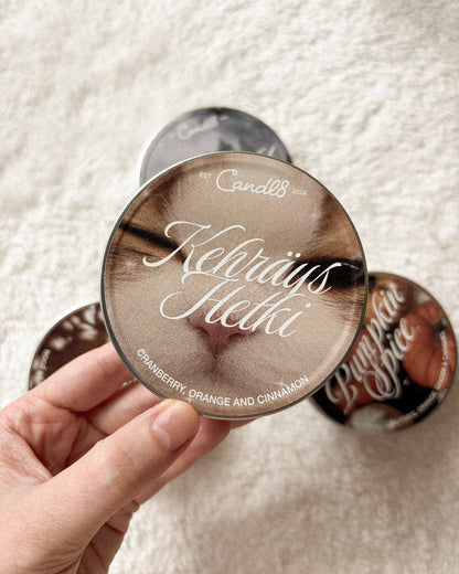 A hand holds a round metal tin of Kehräys Hetki candle, showing the lid printed with a close-up of a cat's face and the scent name 'Kehräys Hetki'. Other matching candle tins are visible on a soft white textured fabric background.