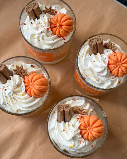 Four glass dessert cups filled with whipped cream and topped with orange pumpkin decorations, cinnamon sticks, star anise, and red sprinkles, arranged on brown paper.