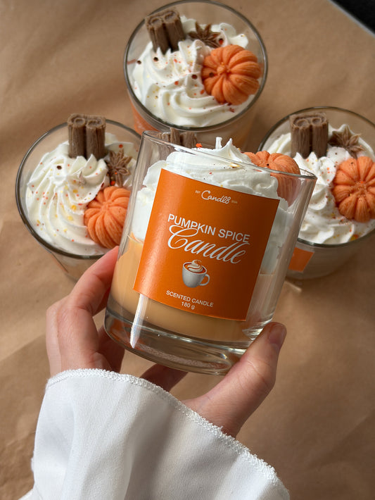 A hand holds a glass jar candle labeled 'Pumpkin Spice Candle' in front of three similar candles topped with whipped cream, orange pumpkin decorations, and cinnamon sticks on a tan surface.