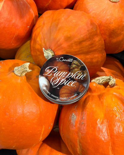 A round tin of Candle's Pumpkin Spice scented candle placed on top of a pile of orange pumpkins. 