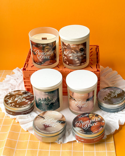 A collection of Candle Couture scented candles in glass jars and tins, featuring labels for Pumpkin Spice, Lammy Ella, Twilight, and Glazed Peach, arranged on a white checkered cloth and orange crate against a solid orange background.