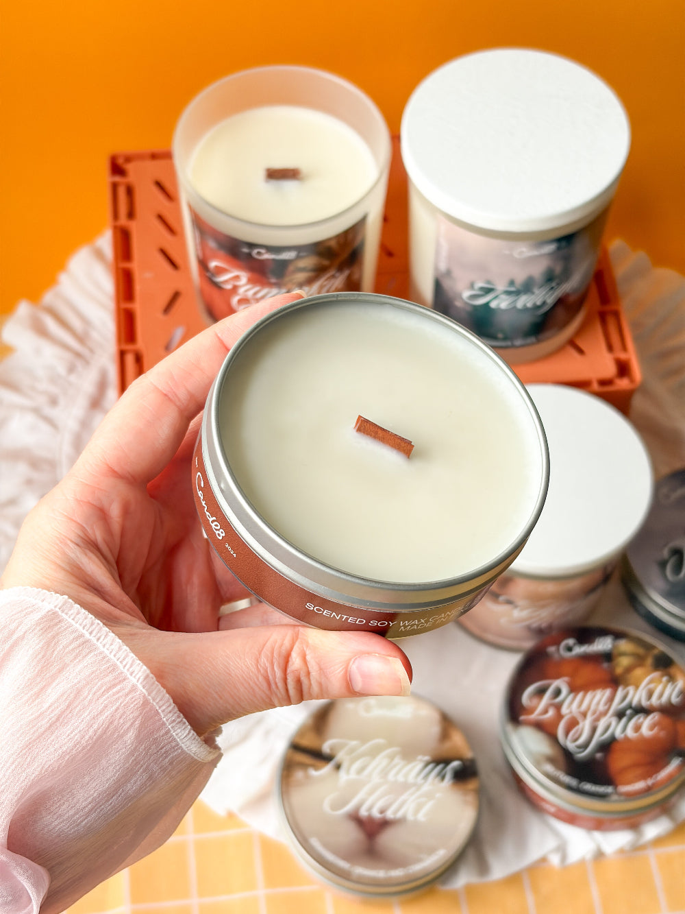 A hand holds a round tin candle with creamy off-white wax and a small wooden wick, with several other scented candles in jars arranged on an orange surface behind it.