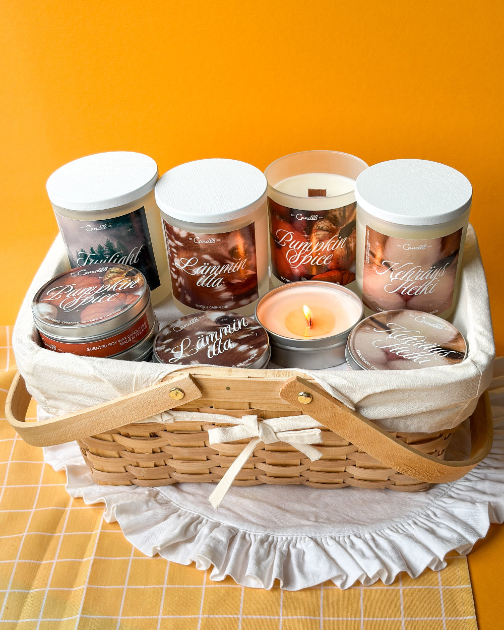 A curated candle gift basket featuring six scented candles in jars and tins, arranged inside a woven wicker basket with a cream fabric liner and wooden handles, set against a solid orange background.