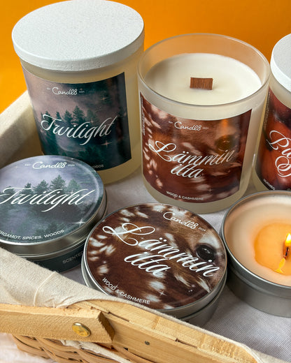 A group of Candle Co. scented candles in glass and tin containers, including Twilight and Luminaria dda variants, arranged in a woven basket with a lit candle glowing on a warm orange background.
