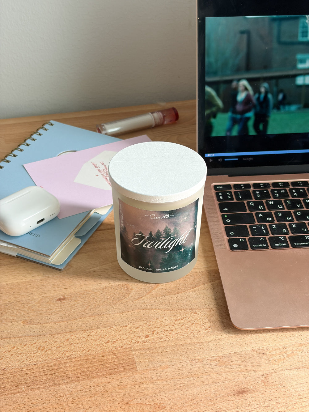 A frosted glass jar candle with a white lid sits on a wooden desk next to an open laptop, a notebook, and other desk accessories. The candle label features a scenic forest graphic with the text 'Fraternal' and the brand name 'Candlelight'.