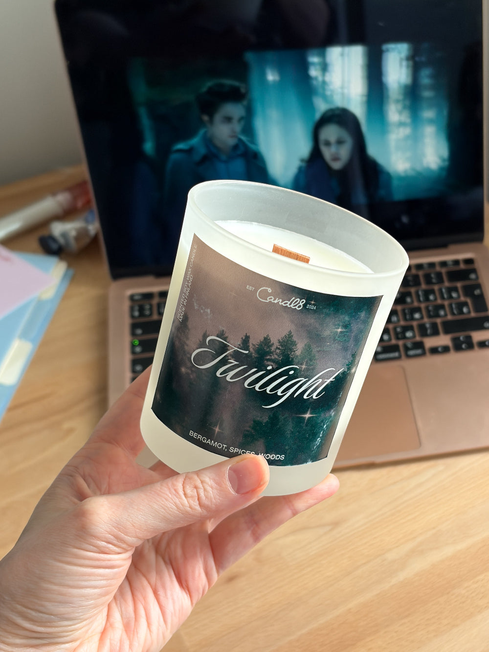 A hand holds a frosted glass candle labeled 'Twilight' in front of an open laptop on a wooden desk.