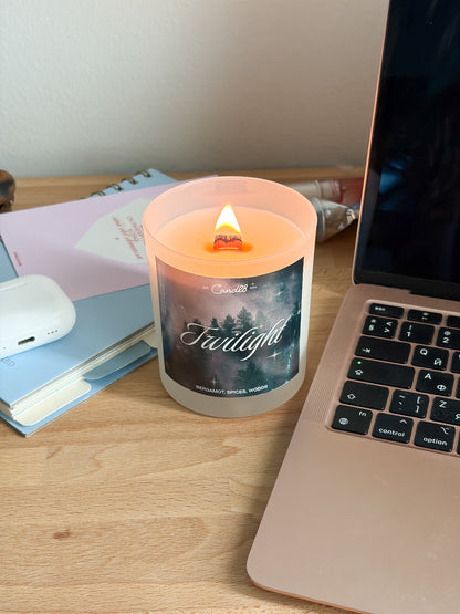 A lit candle in a frosted glass jar sits on a wooden desk next to a laptop, notebooks, and a white earbud case. The candle label reads 'Twilight' and shows a dark forest scene.