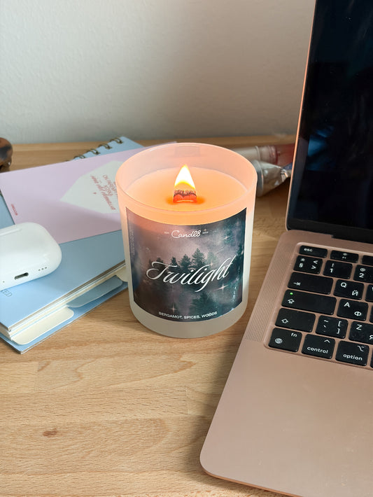 A lit candle in a frosted glass jar sits on a wooden desk next to a laptop, notebooks, and a white earbud case. The candle label reads 'Twilight' and shows a dark forest scene.