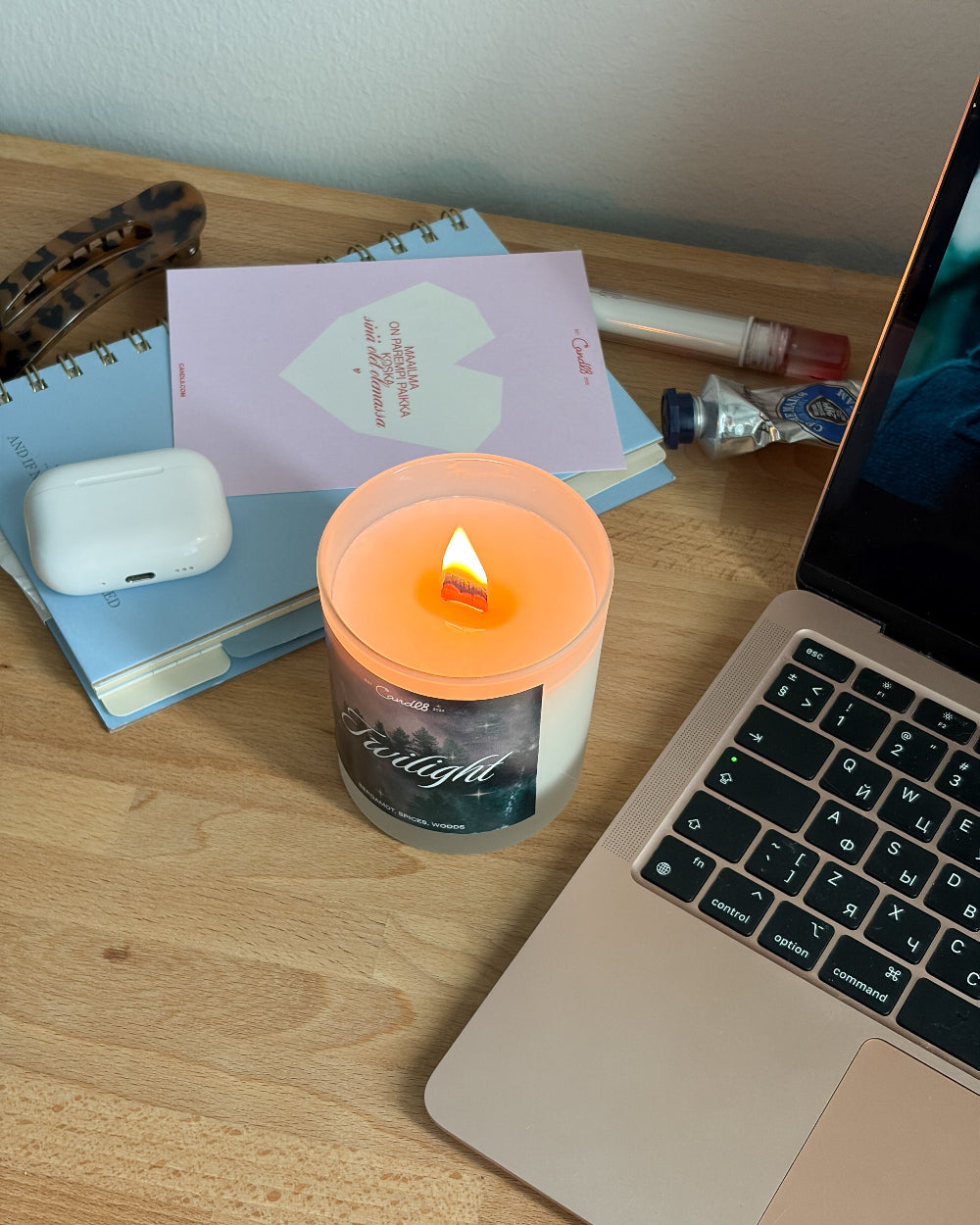 A lit candle in a clear glass jar sits on a wooden desk next to a laptop, notebooks, and other desk accessories. The candle has a warm orange glow and a label that reads 'Twilight'.