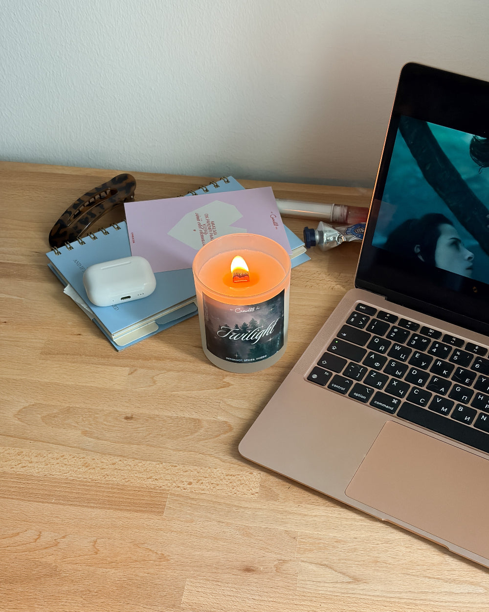 A lit orange candle with a 'Faith' label sits on a light wood desk next to a laptop, notebooks, and a white earbud case.