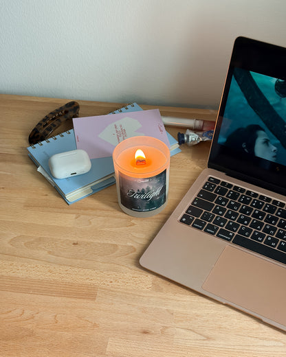 A lit orange candle with a 'Faith' label sits on a light wood desk next to a laptop, notebooks, and a white earbud case.